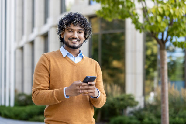 istock Smiling young man using smartphone outdoors in casual autumn attire, standing confidently near modern office building 2174722698