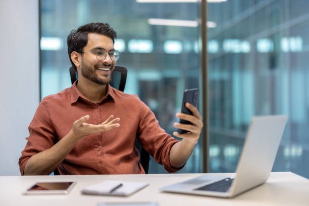 istock Smiling businessman using smartphone for video call at office desk with laptop during work day 2174723663