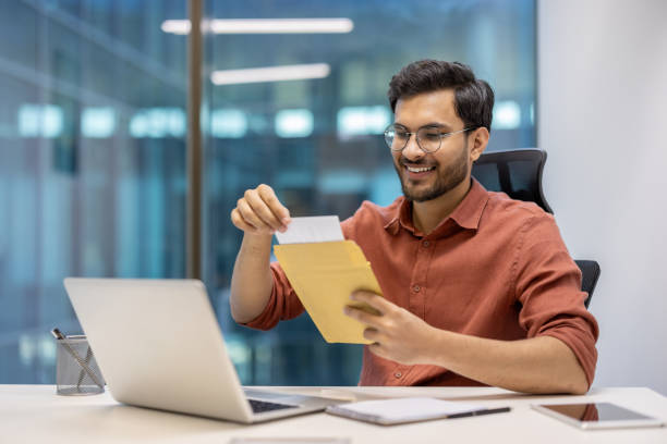 istock Smiling office worker opening envelope while working at desk with laptop and phone in modern workspace 2174723963