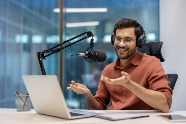 istock Smiling podcast host wearing headphones during online broadcast in modern office setting with laptop and microphone. 2174736181