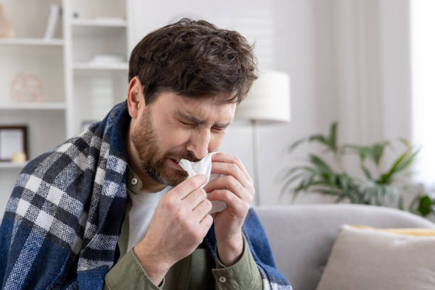 istock Man with cold symptoms sneezing into tissue, wrapped in cozy blanket at home, experiencing seasonal illness 2174739179