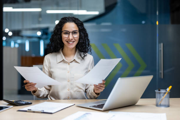 istock Smiling Hispanic businesswoman in modern office holding documents and looking confident with glasses and curly hair 2174739650