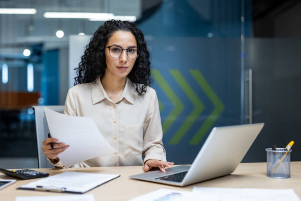 istock Confident businesswoman working in a modern office holding documents and using a laptop for financial analysis and productivity 2174739750