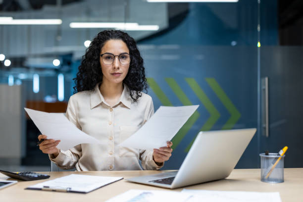 Focused Hispanic businesswoman analyzing documents with thoughtful demeanor in modern office setting