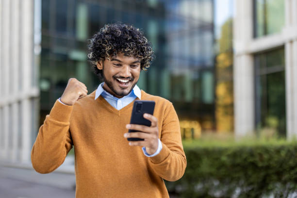 istock Cheerful young man celebrating success while using smartphone outdoors in modern urban setting 2174749612