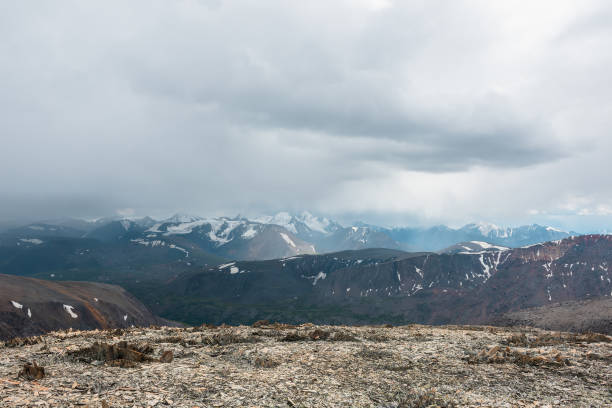 istock Dramatic top view from sunlit stony pass to alpine valley against large mountain range with snow-capped peaks in gray rainy low clouds. Misty snowy mountain tops in grey cloudy sky. Sunlight and rain. 2174749862