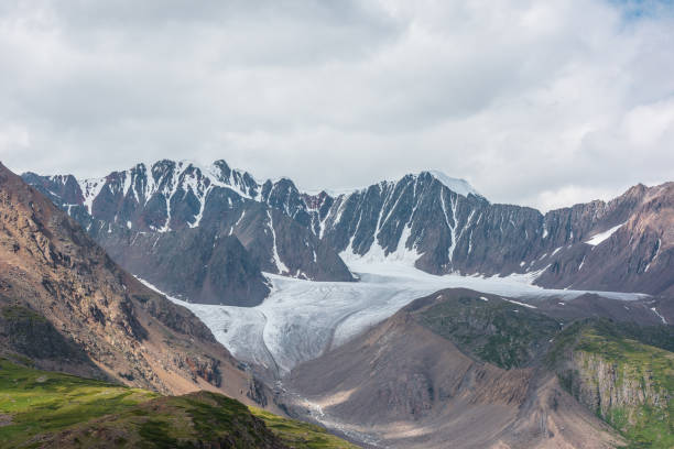 Dramatic landscape with glacier tongue among rocky ridges and sharp snow-capped mountain range under gray cloudy sky. Alpine valley among green hills and rocks in sunlight against ice and sheer crags.