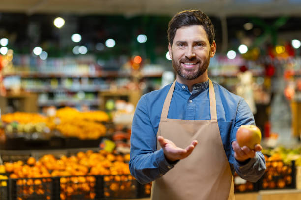 istock Smiling grocery store worker in apron presenting fresh apple in produce section with vibrant fruit in background 2174750148