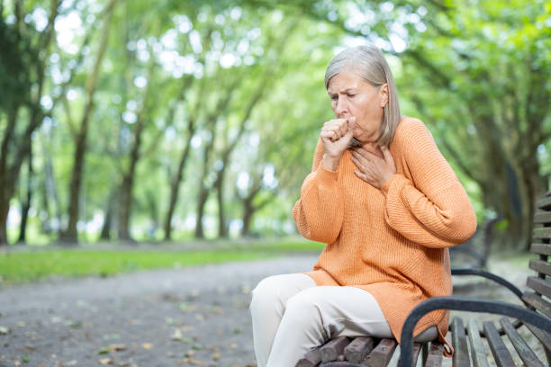 Senior woman experiences difficulty breathing while sitting on park bench, wearing orange sweater in peaceful nature setting, highlighting health issues