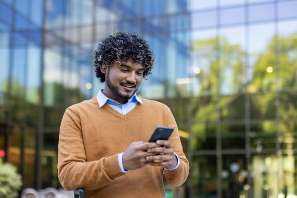 istock Smiling young man using smartphone outside modern office building, wearing casual sweater and blue shirt, enjoying a sunny day 2174751653