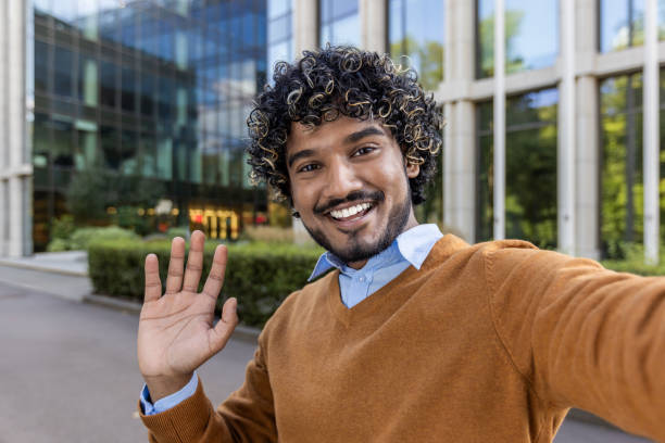 istock Smiling man taking selfie outside modern office building, waving hello to camera, showcasing friendly outdoor environment 2174751971