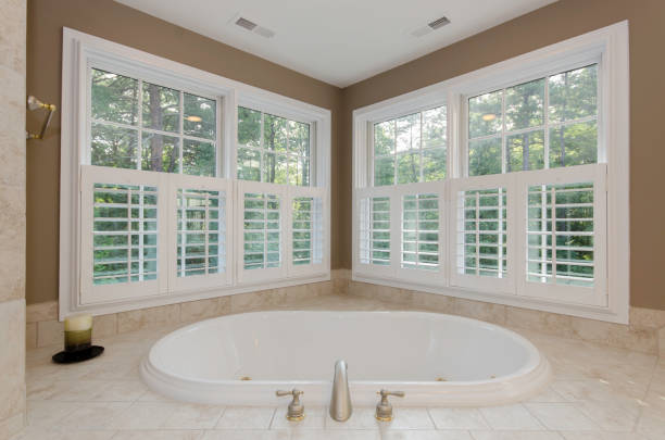 A luxurious bathroom featuring a corner bathtub surrounded by large windows with shutters, inviting natural light and providing a comfortable, relaxing atmosphere.