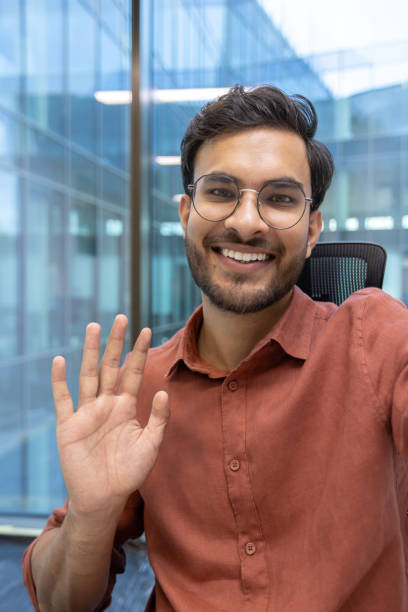 istock Smiling young man waving hello during a video call in a modern office setting, showcasing friendly communication and positive interaction 2174753179