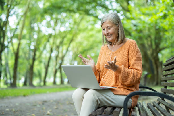 Smiling mature woman using laptop in park enjoying online communication while sitting on bench outdoors
