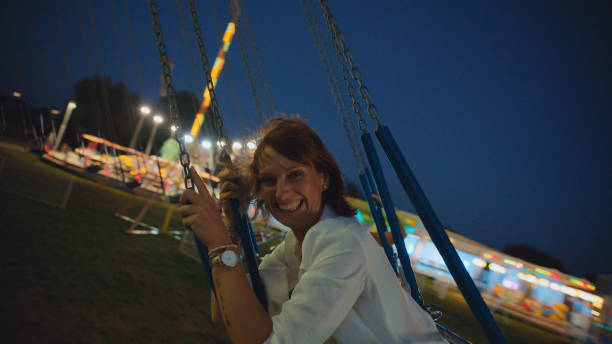 A delighted Caucasian woman enjoys the thrills of a swing carousel ride during a night event, dressed casually with bright lights in the background.