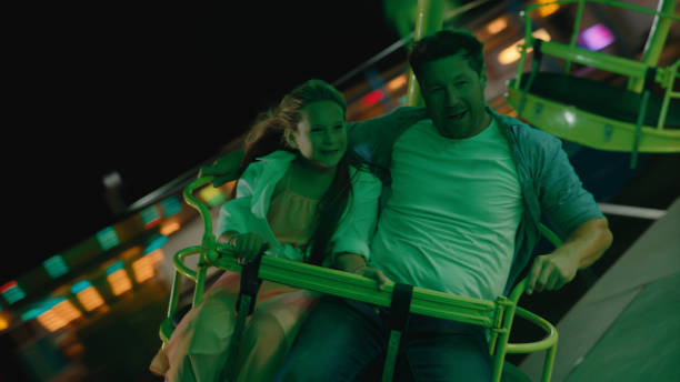 A smiling man and young girl, dressed in casual clothing, delight in the motion of a swing carousel at an amusement park during evening hours.