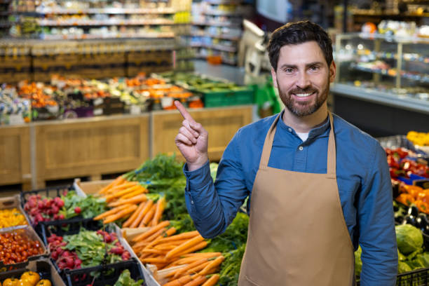 istock Friendly grocery store employee pointing at fresh vegetables in vibrant market setting 2174755929
