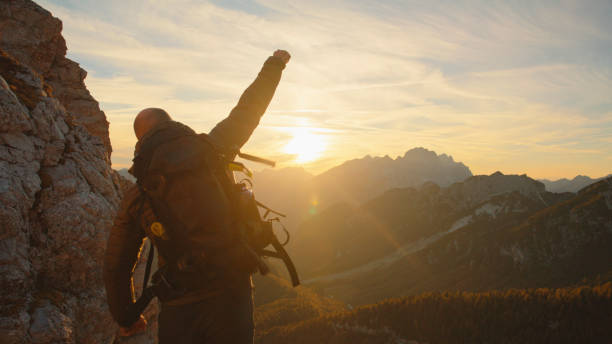 A person in hiking gear triumphantly reaches the peak, celebrating against a backdrop of the setting sun and rugged terrain.