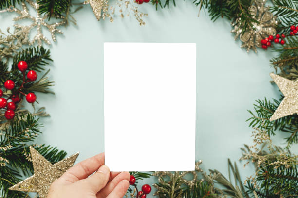 istock Christmas card mock up. Hand holding empty greeting card on blue background with fir branches, red berries and golden ornaments. Merry Christmas. Season greeting postcard template 2174758217