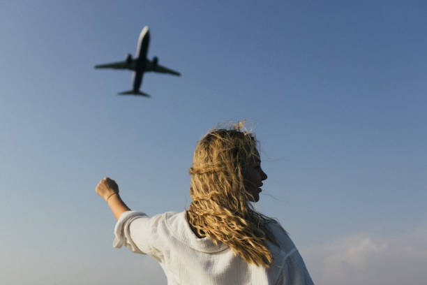 A woman with her arms raised above her head waves at an airplane flying over her.