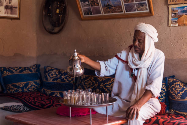 An artisan carefully pours tea into glasses from a silver pot, surrounded by vibrant textiles and a cozy seating area in a traditional Moroccan room.