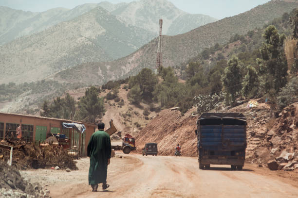 A man walks alongside a dirt road in Morocco's mountainous region, where vehicles navigate the rugged terrain. The landscape features hills, greenery, and a clear blue sky.