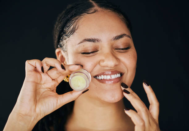 A cheerful woman applies lip balm, enjoying her skincare routine. The black background accentuates her radiant smile and the joy of self-care.