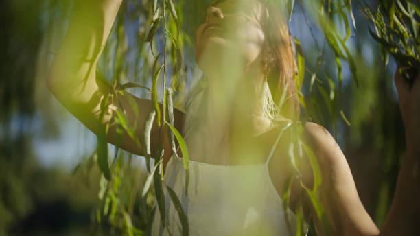 A mature Caucasian woman enjoys a tranquil moment, thoughtfully observing the delicate leaves of a sprawling willow tree, basking in the serene outdoor lighting.