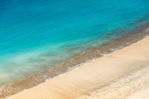 A coastal scene with a clear demarcation between the sandy beach and the ocean. The water transitions from a pale turquoise near the shore to a deeper blue, indicating increasing depth