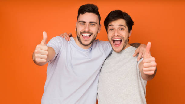 istock Two young men smiling and giving thumbs up against a bright orange background during a cheerful moment of celebration in a casual setting 2174923980