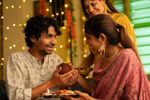 Woman looking at sister giving religious offerings to brother during Bhai Dooj Smiling young woman looking at sister giving coconut to smiling brother while celebrating Bhai Dooj festival in living room at home bhai tika stock pictures, royalty-free photos & images