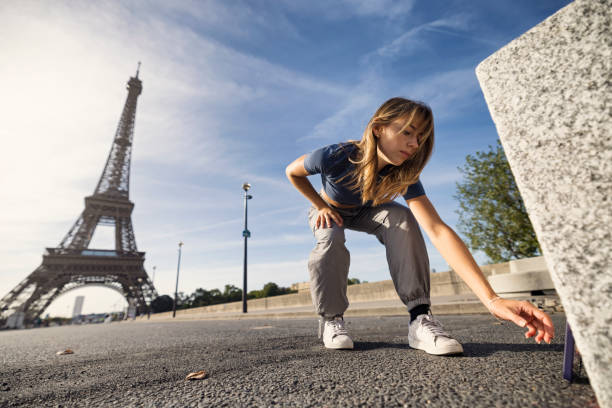 Young woman is crouching on the ground near the Eiffel Tower, carefully adjusting a smartphone placed against a stone block. The tower stands tall in the background, while she concentrates on setting the perfect angle. \nShot With Canon R5