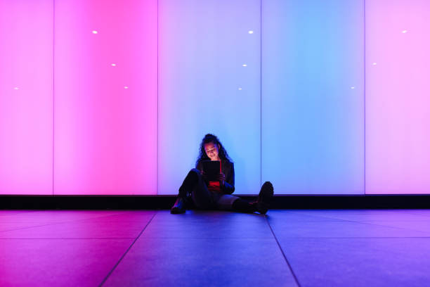 istock Young woman using digital tablet sitting on floor with neon lights background 2174951483