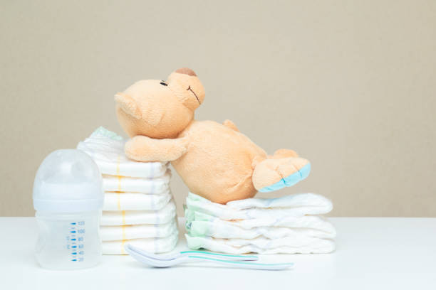 istock Stack of diapers with teddy bear toy lying on white table, bottle for milk and spoons. set for baby shower, copy space 2174954390