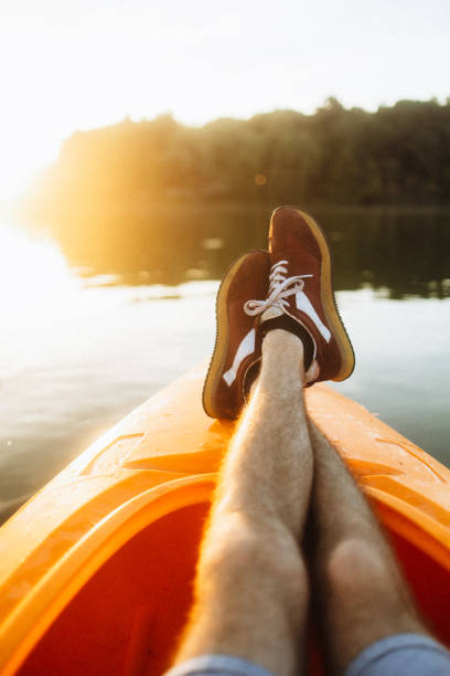 A peaceful scene of a man stretching his legs while relaxing on a kayak at sunset. The warm, golden light of the setting sun reflects off the calm water, creating a tranquil and serene atmosphere