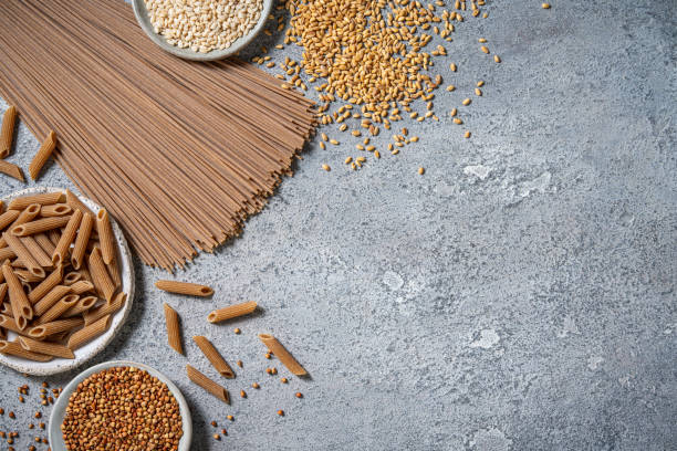 A collection of high-fiber vegan foods is displayed on a gray kitchen table, featuring whole grain pasta, cereals, and various seeds, showcasing nutritious dietary options.