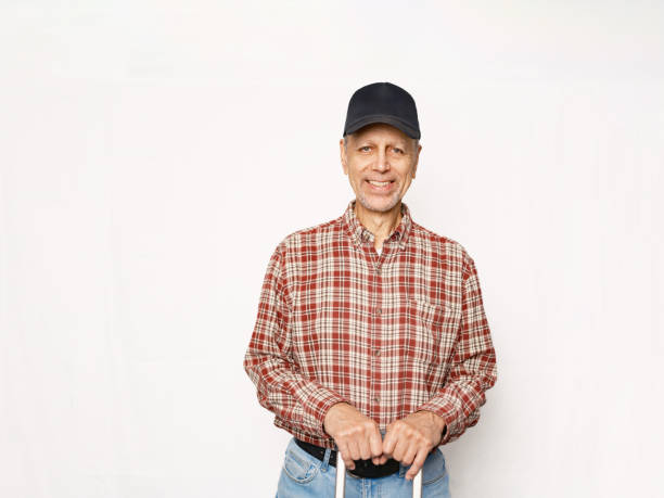 Senior Caucasian man against white backdrop. He is dressed in casual clothes and ball cap..