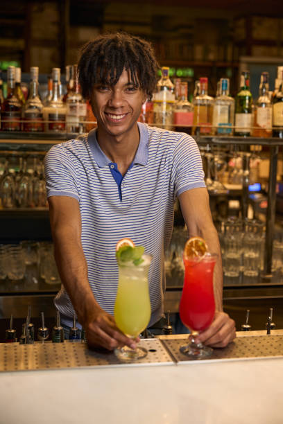 Young man standing behind the bar counter, smiling with three colorful cocktails placed in front of him