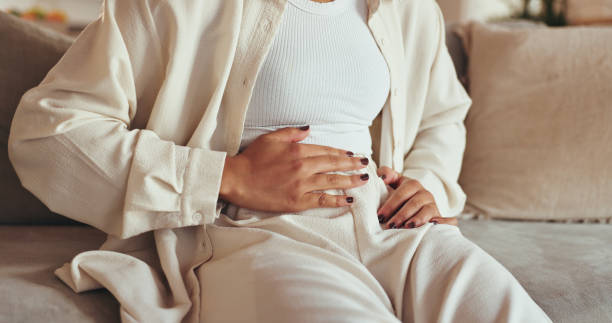 woman, hands and stomach pain of menstruation on sofa with period cramps, bloating or pms in home. person, abdomen and endometriosis with constipation, nausea and digestion problem on couch in lounge - pijn fotos stockfoto's en -beelden