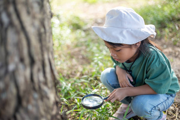 A young girl is looking at a tree with a magnifying glass. She is wearing a white hat and a green shirt