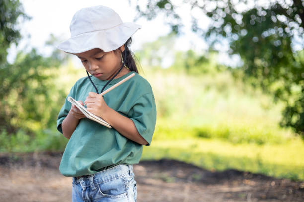 A young girl is looking at a tablet while wearing a white hat. She is writing in a notebook