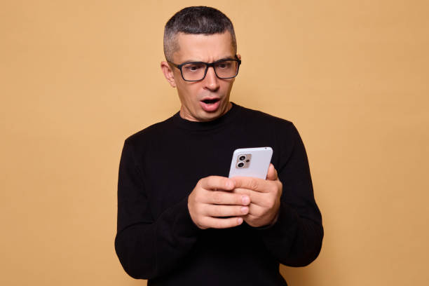 istock Shocked Caucasian man wearing black jumper and spectacles focused on his smartphone while checking a social media app with confused expression isolated over beige background 2175132663
