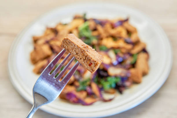 A close-up shot of a fork holding a piece of tofu above a plate of colorful stir-fried vegetables, showcasing a delicious vegan meal. stock photo