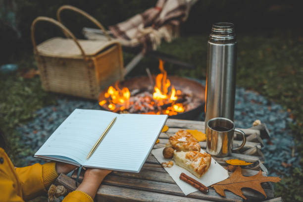istock Person writing in a notebook next to a bonfire during autumn. Outdoor cozy setting with fallen leaves, snacks, hot drink, and the warmth of the fire, capturing the essence of a peaceful autumn day 2175153216