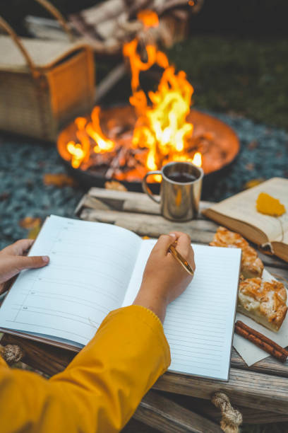 istock Person writing in a notebook next to a bonfire during autumn. Outdoor cozy setting with fallen leaves, snacks, hot drink, and the warmth of the fire, capturing the essence of a peaceful autumn day 2175153223
