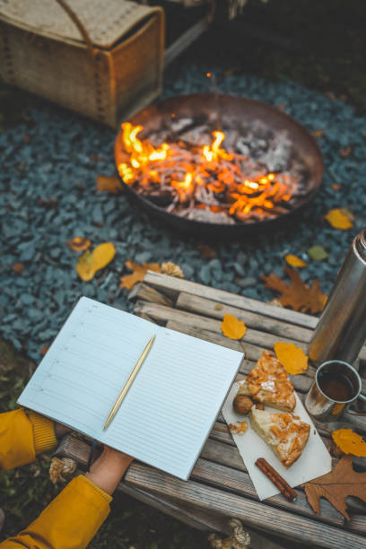 istock Person writing in a notebook next to a bonfire during autumn. Outdoor cozy setting with fallen leaves, snacks, hot drink, and the warmth of the fire, capturing the essence of a peaceful autumn day 2175153233