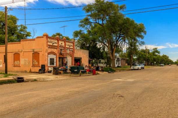Las Animas, Colorado, Bent County, USA - May 31, 2024: Rustic brick building with 'Hunting' and 'Fishing' signs is a small thrift store displays vintage outdoor equipment and furniture for sale. Tree-lined streets, residential houses, and utility poles create a peaceful small-town atmosphere. Bright blue sky with puffy white clouds overhead.