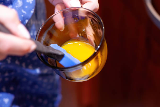 A person uses a blue spatula to mix egg yolk in a glass bowl. The image captures the action in a close-up, highlighting the vibrant yellow color and smooth texture of the yolk.