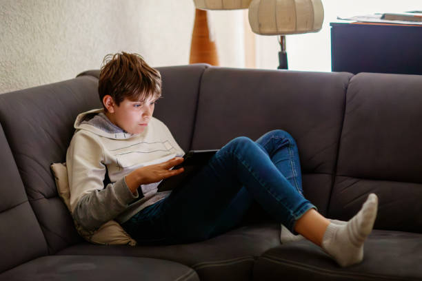 istock Teenager boy lounging comfortably on a grey couch while using a tablet. Child is relaxed, holding the tablet with both hands, and appears to be engaged in what he is watching or reading, or gaming 2175169856