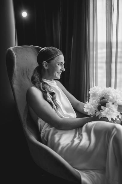 Black and white portrait of young bride sitting in armchair, holding bouquet of flowers and looking through window before wedding ceremony